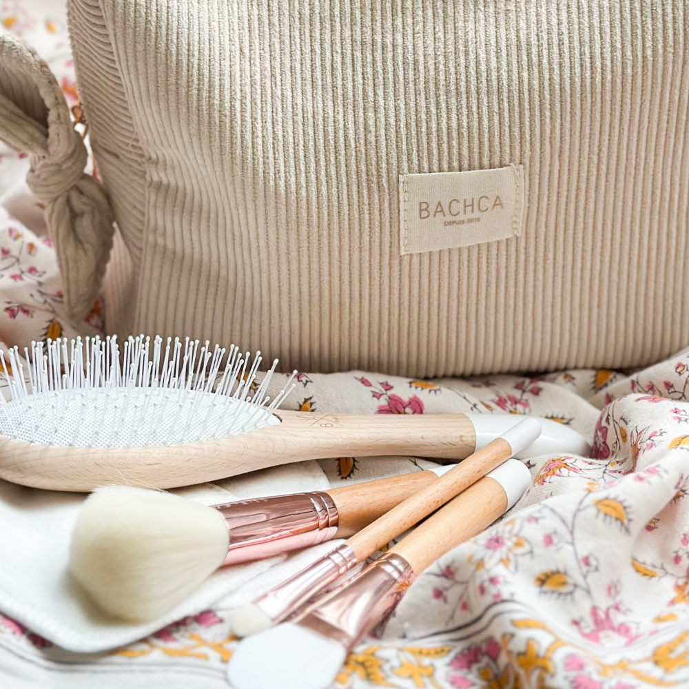 Zoom sur une trousse de toilette en velours de la marque Bachca, des accessoires beauté de la même marque sont disposés devant la trousse sur un linge fleuri.