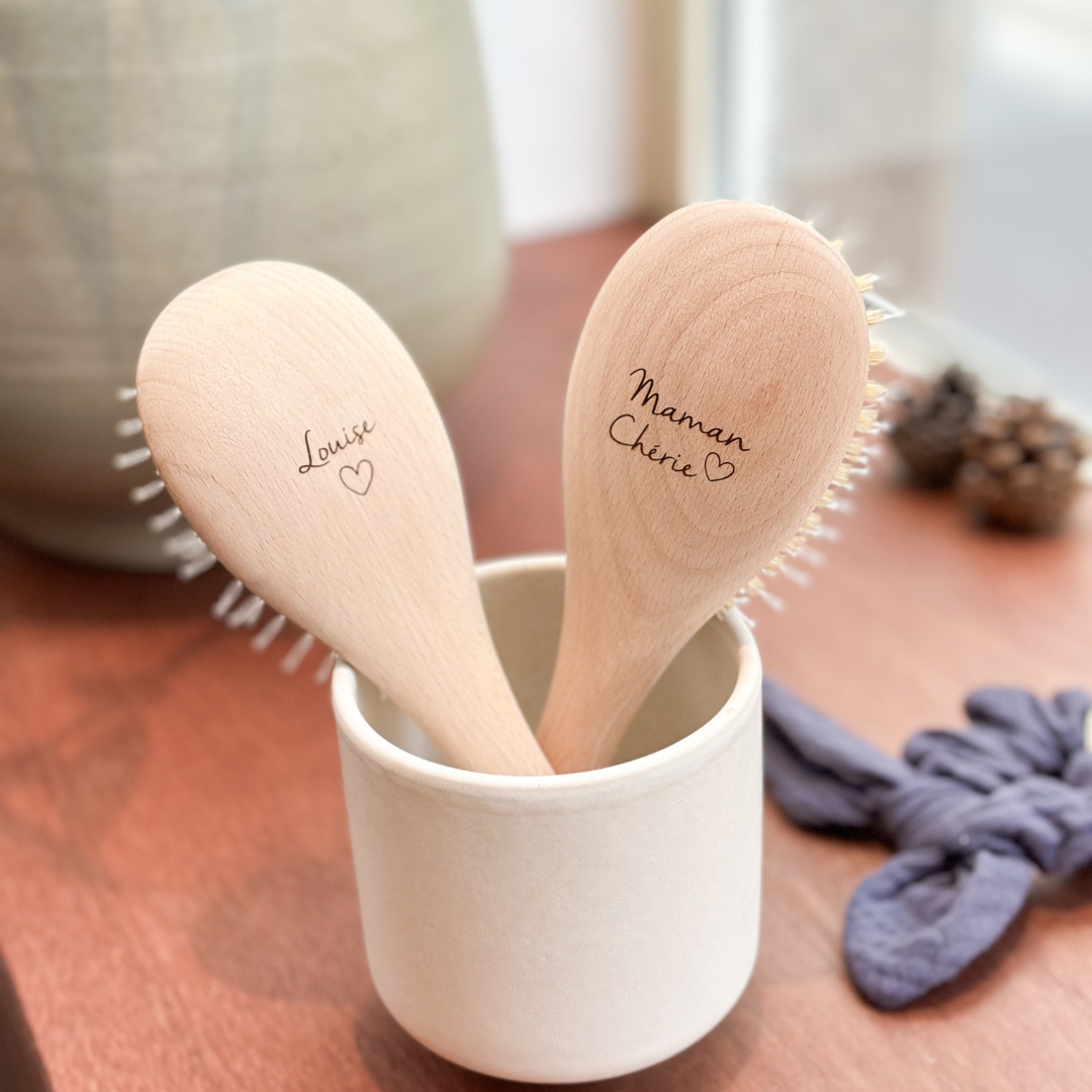 Deux brosses à cheveux Bachca de dos dans un pot. Gravées chacune avec "Louise ♥" et "Maman Chérie ♥", sur un meuble en bois.