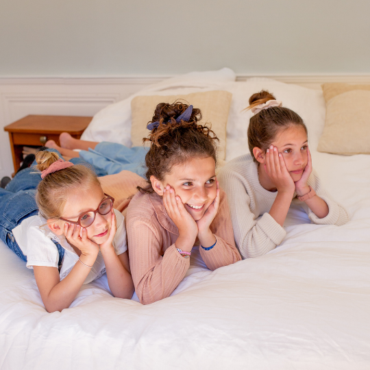 Trois filles allongées sur un lit, les mains posées sur les joues, portant des chouchous en gaze de coton Bachca dans les cheveux attachés en chignon.