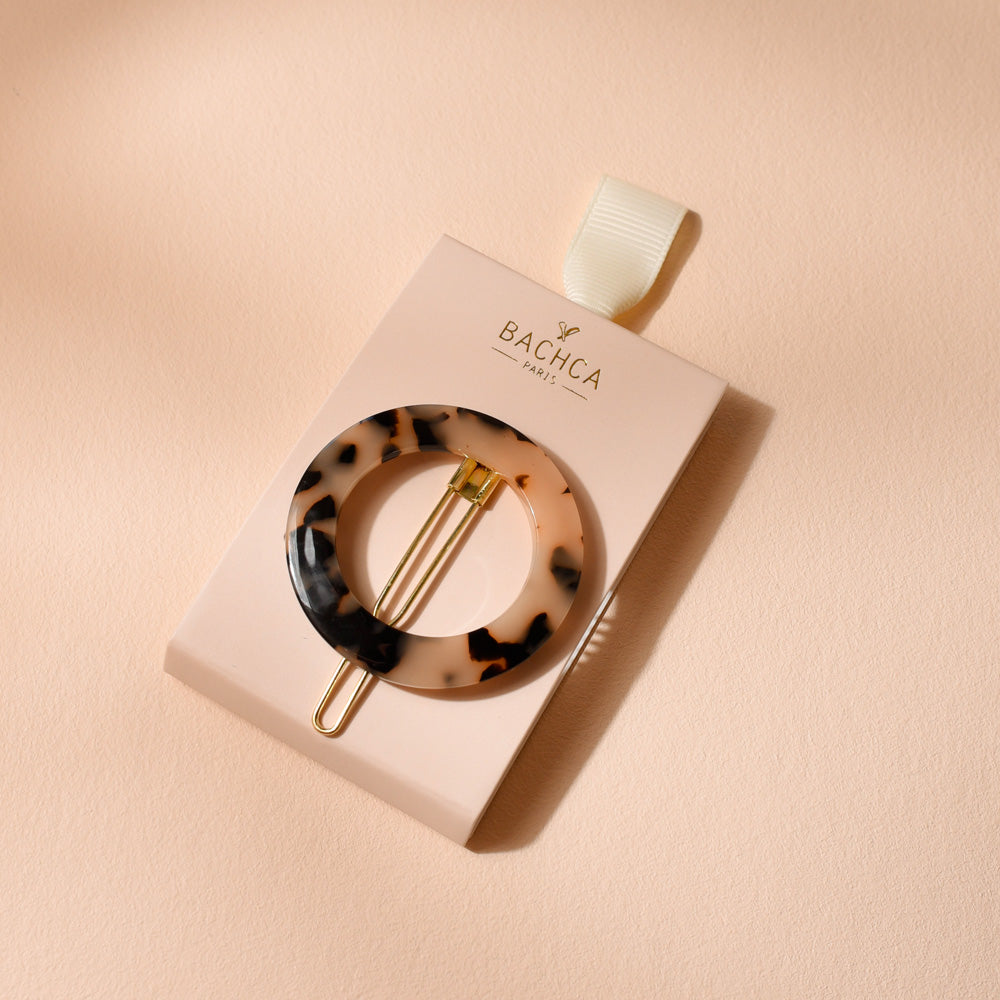 Barrette ronde Bachca en acétate motif écaille avec fermoir doré, présentée sur son support cartonné rose poudré, photographiée sur une surface beige.