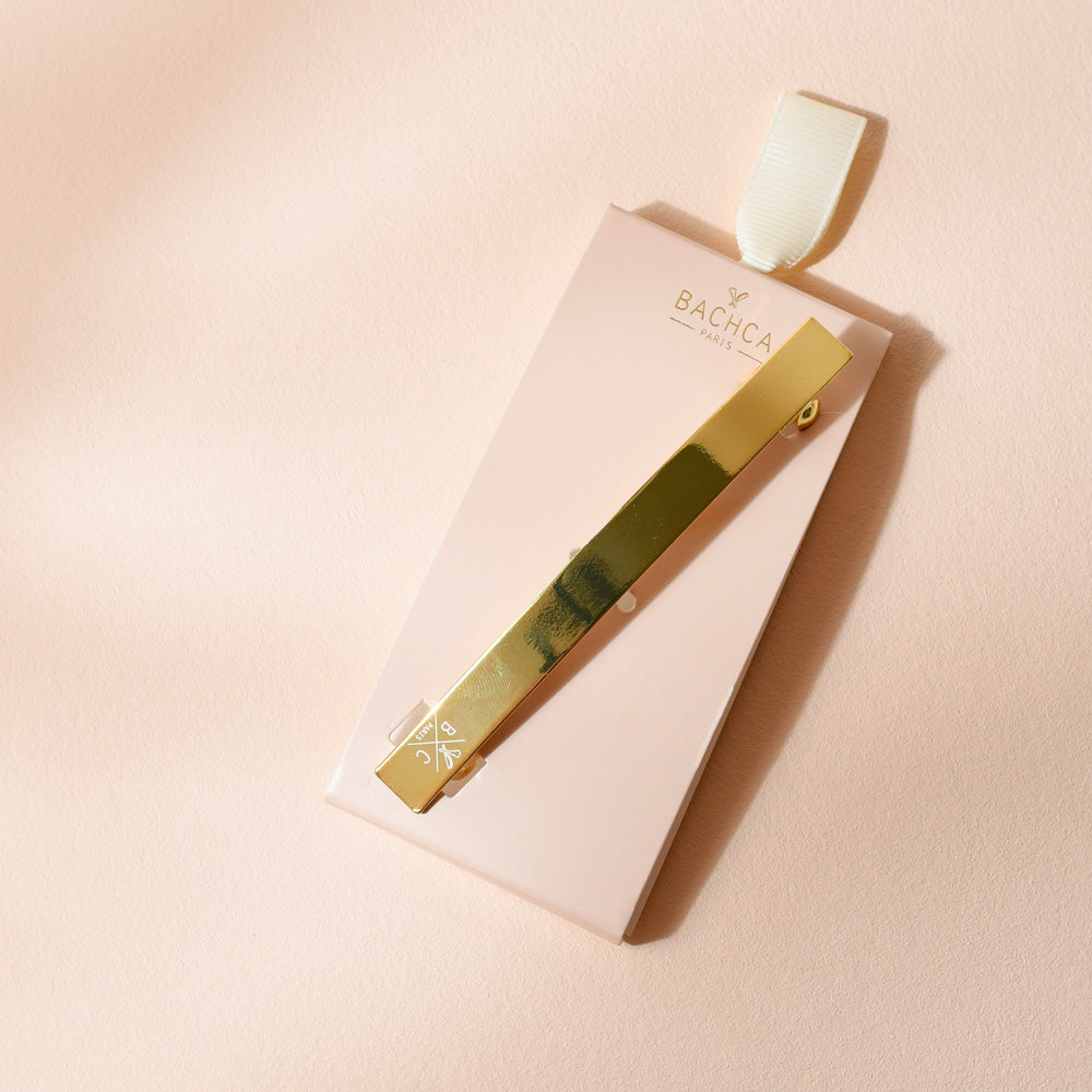 Barrette longue Bachca en métal doré, présentée sur son support cartonné rose poudré, photographiée sur une surface beige.