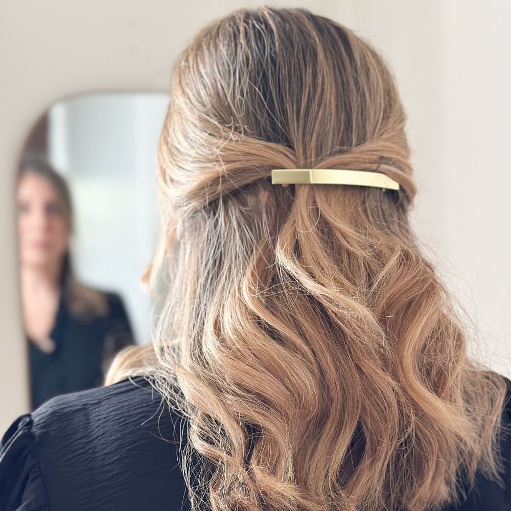 Cheveux blonds ondulés retenus en demi-queue par une barrette longue dorée minimaliste Bachca.