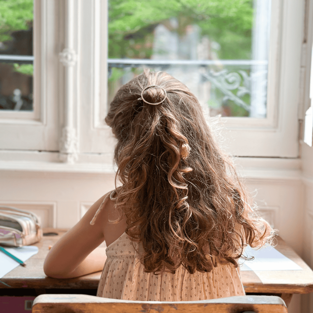 Petite fille de dos qui dessine. Elle porte dans ses cheveux une barrette ronde dorée de la marque Bachca.