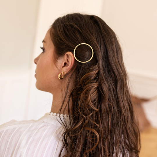 Femme aux cheveux bruns ondulés portant une barrette ronde dorée Bachca sur le côté, coiffure élégante et raffinée.