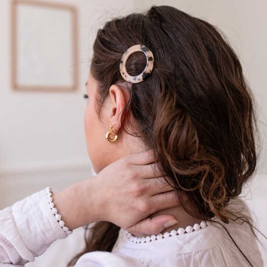 Femme aux cheveux bruns ondulés portant une barrette ronde écaille Bachca sur le côté, coiffure élégante et raffinée.