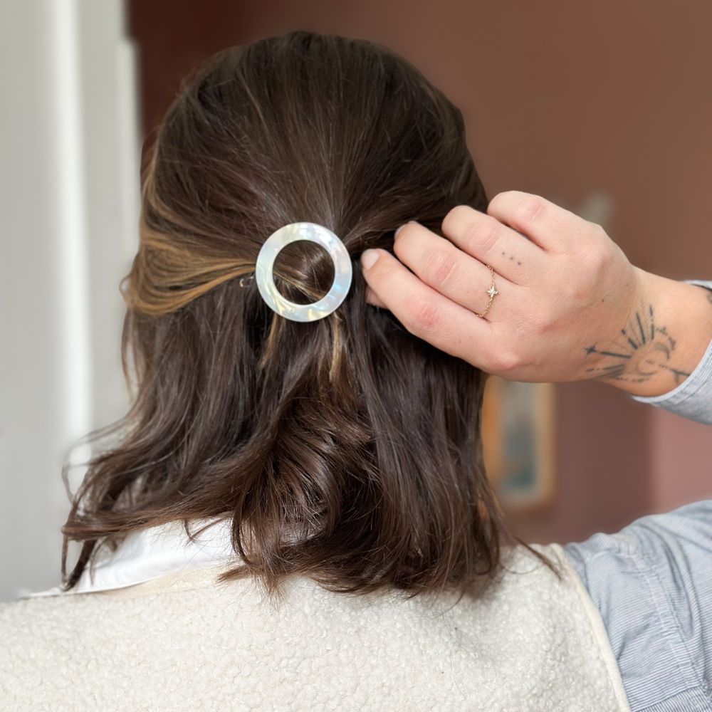 Cheveux bruns attachés en demi-queue avec barrette ronde nacrée Bachca, mise en beauté naturelle et élégante au fini irisé.
