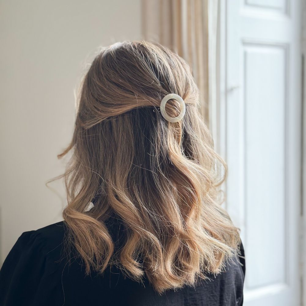 Cheveux ondulés attachés en demi-queue avec barrette ronde nacrée Bachca, coiffure naturelle et lumineuse devant une fenêtre.