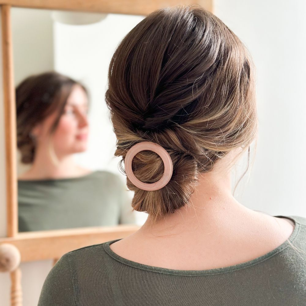 Femme coiffée d’un chignon bas, ornée d’une barrette ronde rose dragée Bachca devant un miroir.