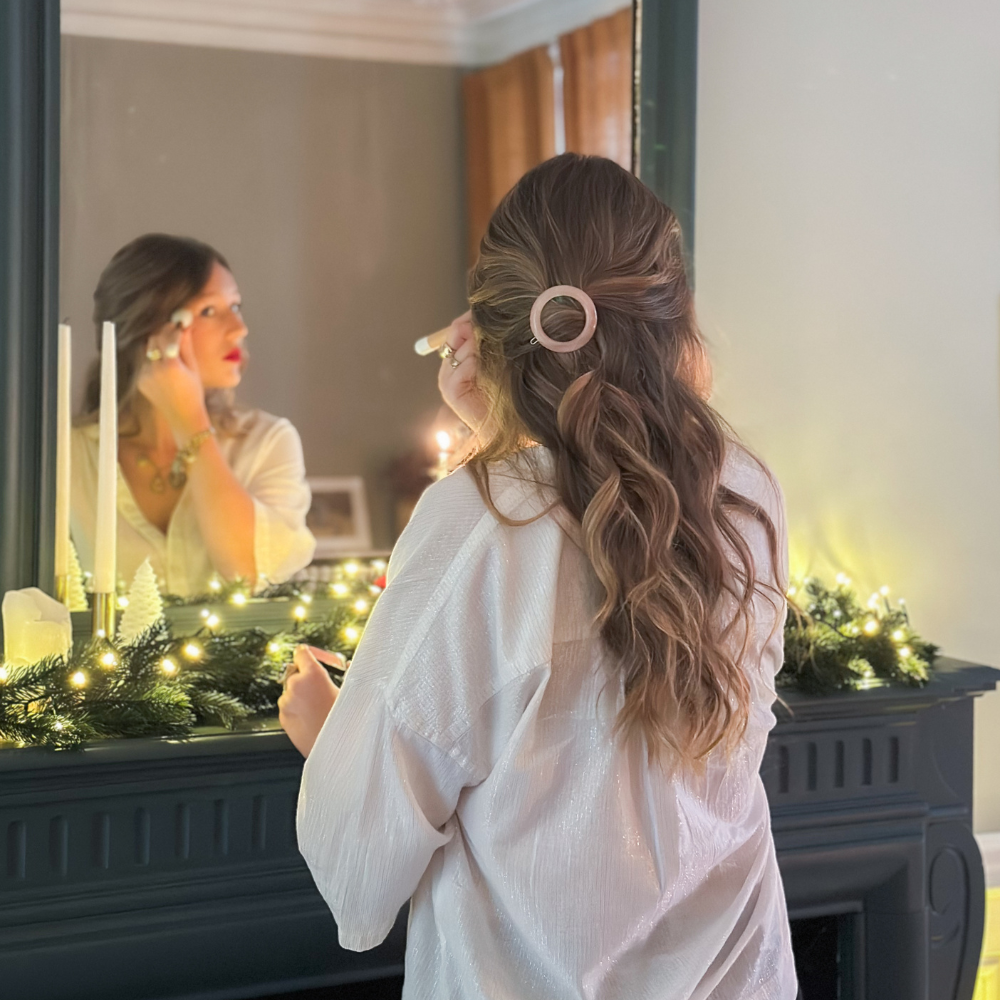Femme qui se maquille face à un miroir, elle à les cheveux longs blonds, elle porte une barrette ronde rose pâle de la marque Bachca dans les cheveux.