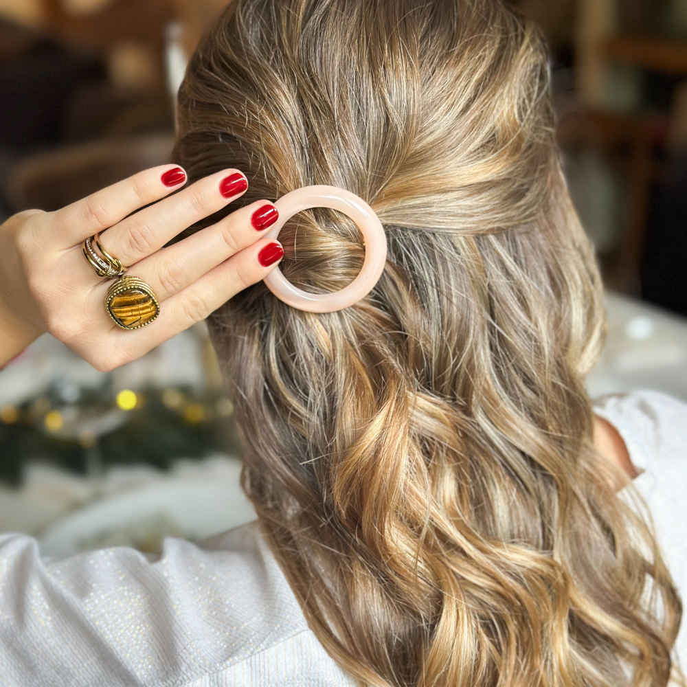Une femme de dos avec de longs cheveux blonds, elle porte une demi-queue attachée avec une barrette ronde rose pâle de la marque Bachca.