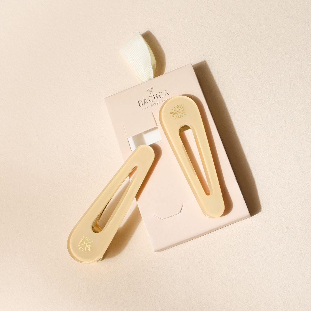 Lot de deux barrettes plates Bachca en acétate ivoire avec fermoir doré, présentées sur leur emballage rose poudré, posées sur fond beige.