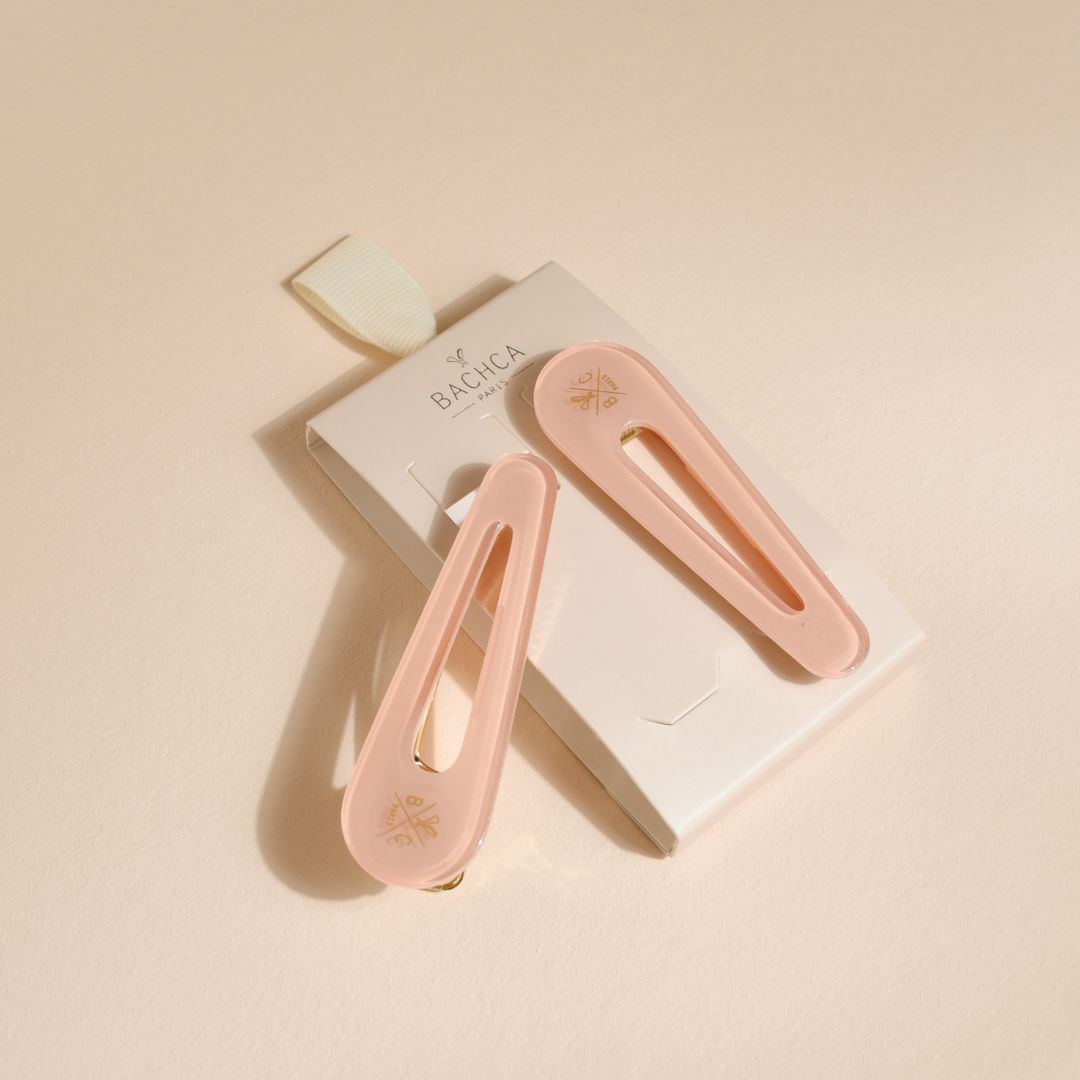Lot de deux barrettes plates Bachca en acétate rose dragée avec détails dorés, présentées sur leur emballage élégant, sur fond beige.