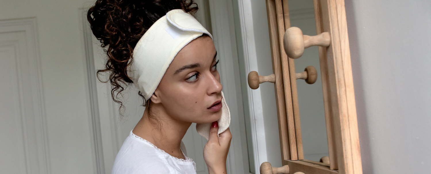 Jeune femme se démaquillant face à un miroir, ses cheveux sont protégés à l'aide d'un bandeau de soin pour les cheveux de la marque Bachca.