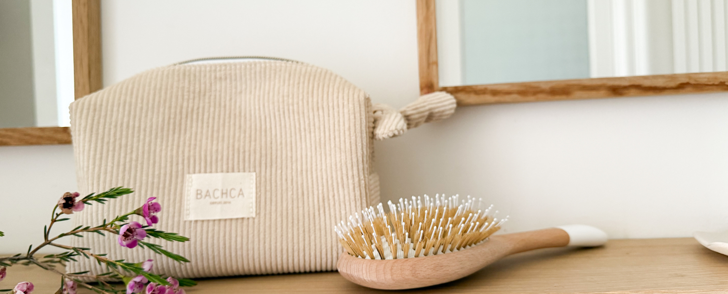 Photo lifestyle de la trousse maquillage en velours côtelé de la marque Bachca sur le rebords d'une étagère de salle de bain accompagnée d'une brosse à cheveux démêlage et brillance.