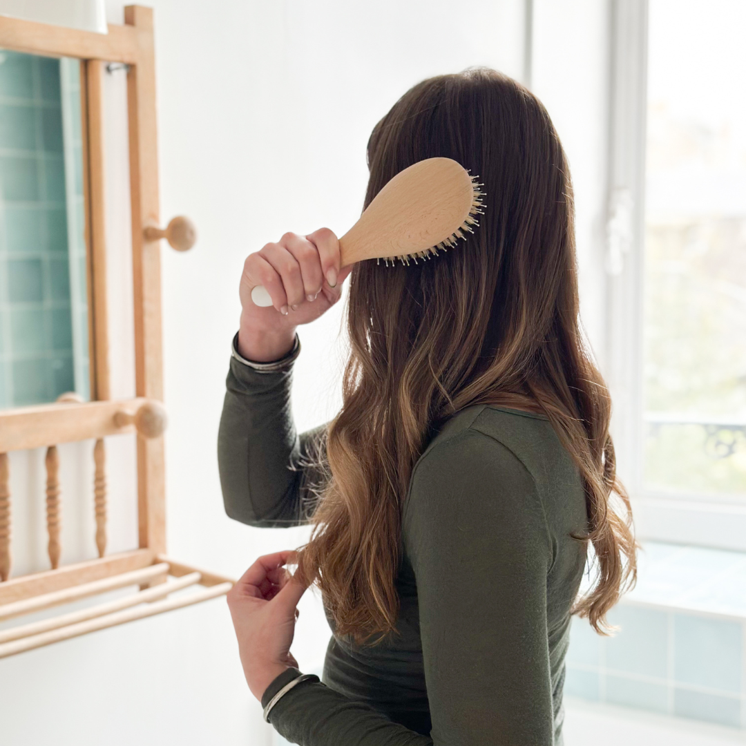 Femme se brossant les cheveux avec une brosse démêlante Bachca en bois, devant un miroir mural dans une salle de bain lumineuse.