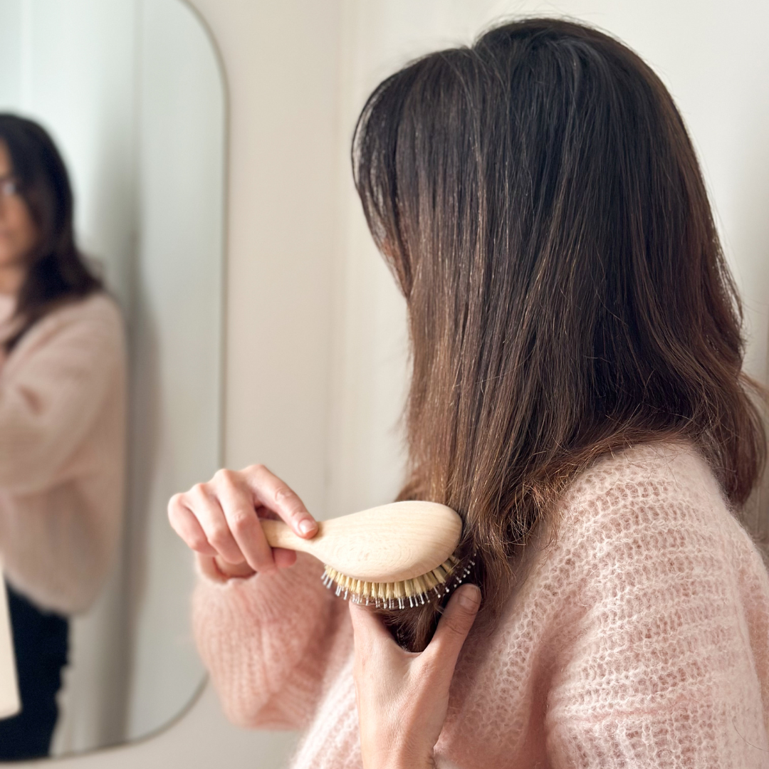 Femme de dos en pull rose se brossant les longueurs avec une brosse Bachca en bois devant un miroir.