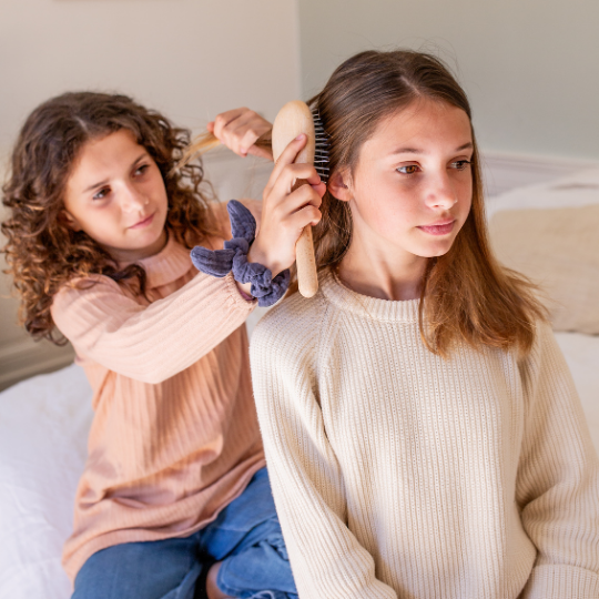 Fillette coiffant les cheveux d’une autre avec une brosse démêlante Bachca en bois, assises sur un lit dans une chambre lumineuse.