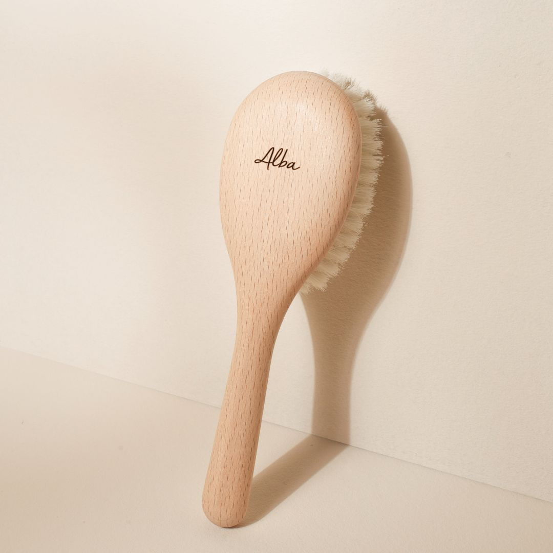Brosse à cheveux Bachca pour bébé, en bois de hêtre brut, gravée du prénom « Alba » sur le dos, avec des poils de chèvre très doux, posée verticalement contre un mur beige.