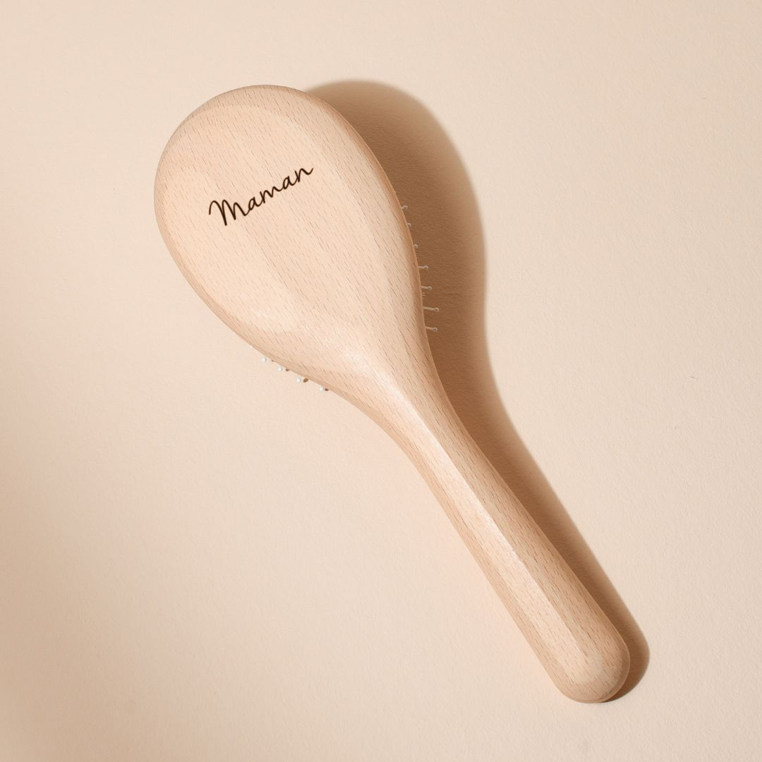 Brosse à cheveux Bachca en bois de hêtre naturel, vue de dos, avec le mot « Maman » gravé délicatement sur le dos du manche, posée sur un fond beige poudré.