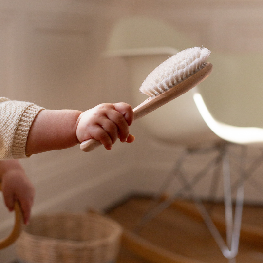 Main d’un bébé tenant une brosse à cheveux Bachca en bois de hêtre brut avec des poils ultra-doux, dans un intérieur chaleureux avec sol en bois et chaise design en arrière-plan.