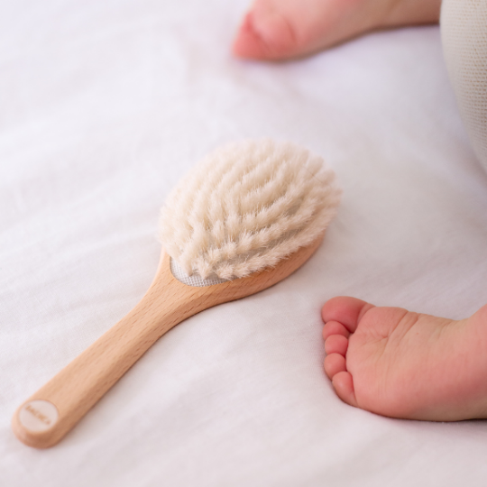 Brosse à cheveux pour bébé Bachca, posée sur un drap blanc près des pieds d’un nourrisson, avec des poils très doux et un manche en bois de hêtre brut, idéale pour coiffer en douceur dès la naissance.