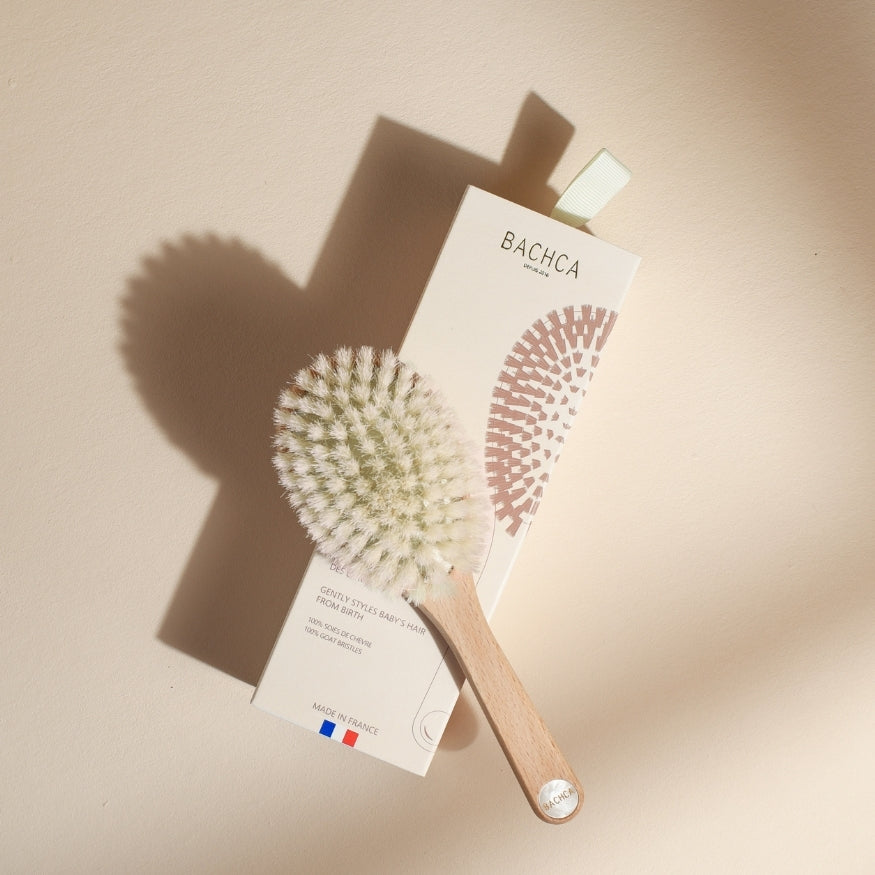 Brosse bébé Bachca posée en diagonale sur son étui crème, avec manche en bois clair et poils blancs très doux, éclairée par une lumière naturelle sur fond beige poudré.