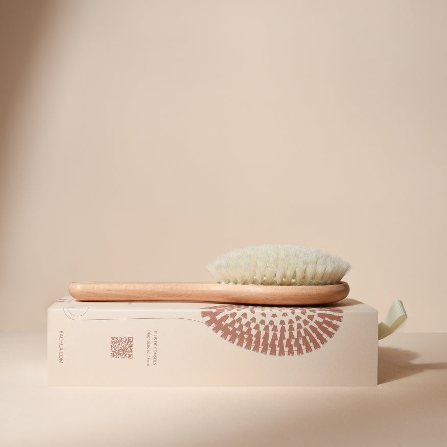 Brosse à cheveux Bachca pour bébé posée sur son étui, avec manche en bois naturel et poils blancs ultra doux, photographiée sur fond beige pour un rendu épuré et apaisant.