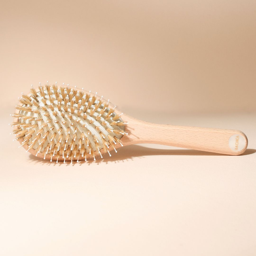 Brosse Ă cheveux Bachca posĂ©e Ă plat, vue sur les picots en bois et nylon, manche en bois de hĂȘtre brut avec pastille nacrĂ©e au bout, sur fond beige uniforme.