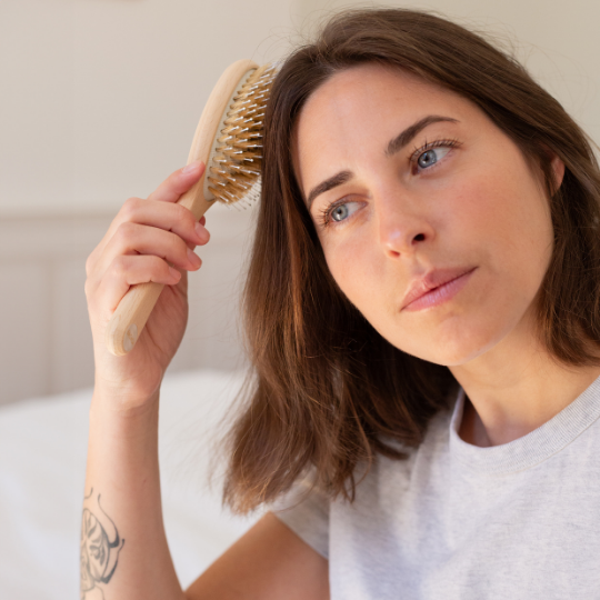 Femme se brossant les cheveux avec une brosse à cheveux Bachca, en bois de hêtre naturel, dotée de picots en nylon et de soies. Elle est assise dans une pièce lumineuse, vêtue d’un t-shirt gris clair.