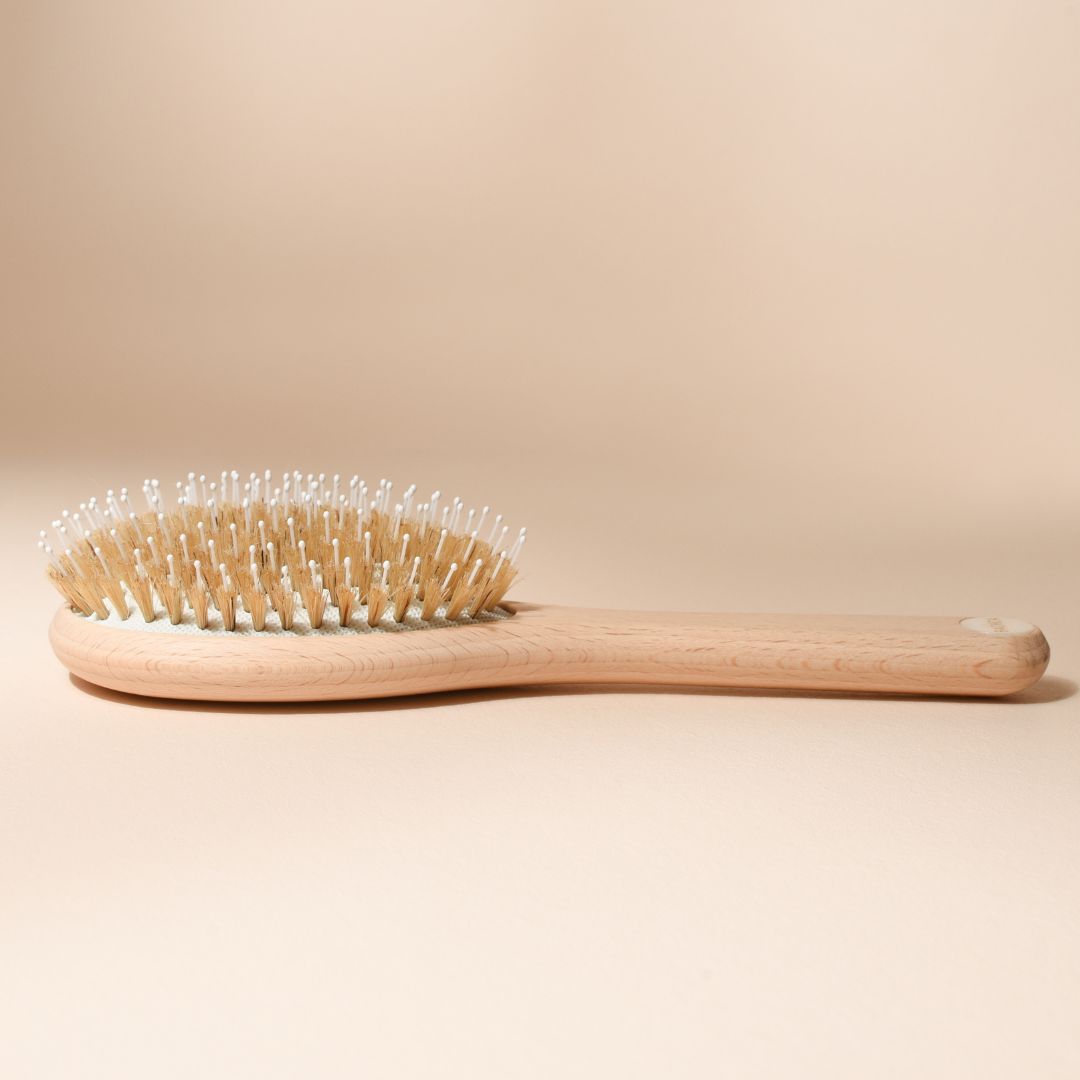 Brosse à cheveux Bachca posée à plat sur fond beige, en vue de profil. Son manche en bois de hêtre naturel est prolongé par un coussin crème garni de picots en nylon et poils de sanglier, avec une pastille nacrée au bout du manche.