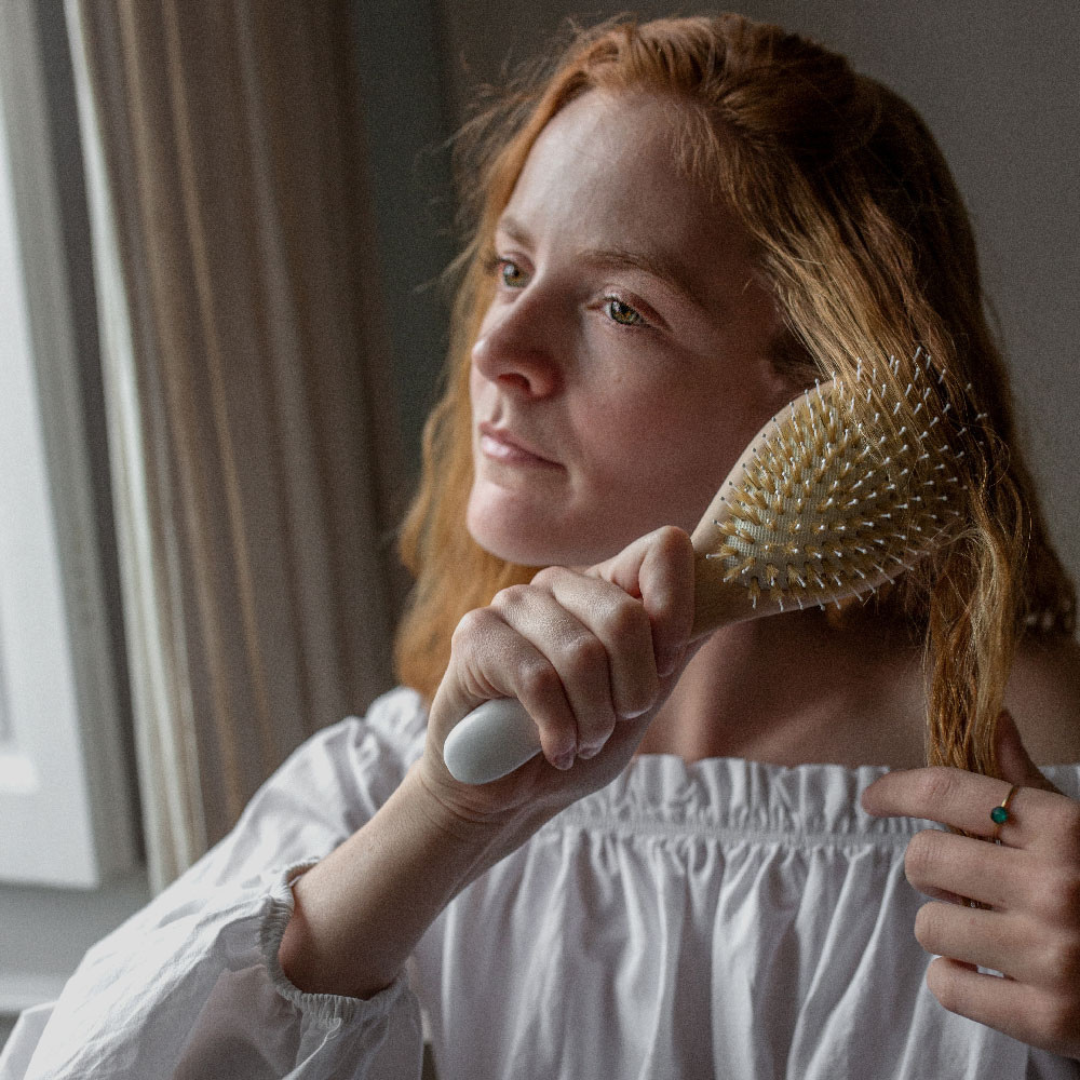 Femme se brossant les cheveux avec une brosse Bachca en bois naturel, près d’une fenêtre, dans une lumière douce et intimiste.