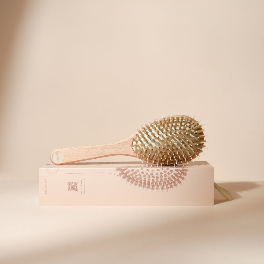 Brosse à cheveux Bachca photographiée de profil, posée sur son coffret illustré, avec manche en bois clair et coussin garni de picots et poils mixtes, sur un fond beige doux.
