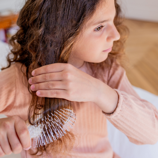 Jeune fille aux cheveux bouclés se brossant les longueurs avec la brosse démêlante et volume Bachca en bois clair, vêtue d’un haut rose poudré.