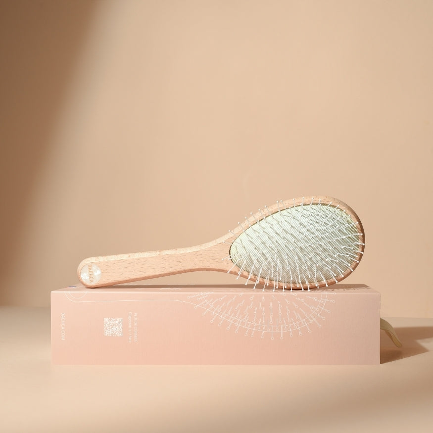 Brosse démêlante universelle Bachca vue de profil, posée sur son coffret rose poudré illustré, avec manche en bois de hêtre clair et coussin garni de picots fins, sur fond beige neutre.