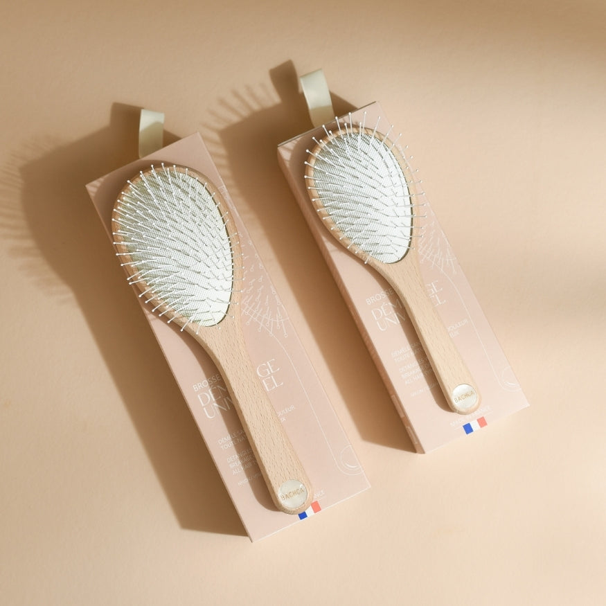 Duo de brosses démêlantes universelles Bachca présentées dans leurs coffrets rose poudré, avec manche en bois clair et coussin à picots blancs, disposées à plat sur fond beige avec une ombre marquée.