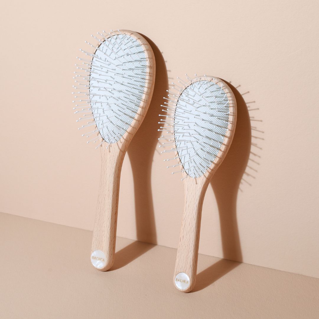 Deux brosses démêlantes universelles Bachca, formats grand et petit, en bois de hêtre brut avec picots nylon blancs, posées debout contre un mur beige.