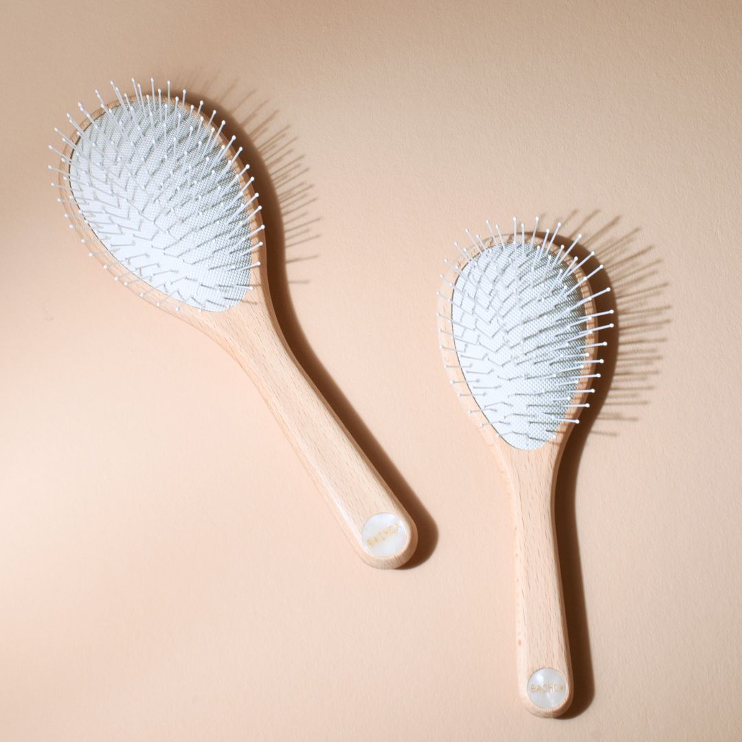 Deux brosses démêlantes universelles Bachca, petit et grand format, en bois de hêtre brut avec picots nylon blancs, posées à plat sur un fond beige.