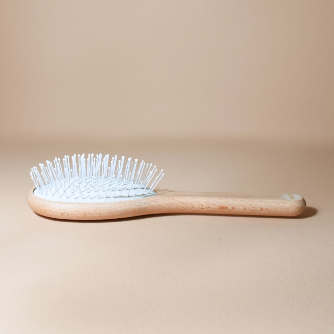 Brosse démêlante universelle Bachca petit format en bois de hêtre brut, vue de côté, avec des picots nylon blancs arrondis, posée sur un fond beige.