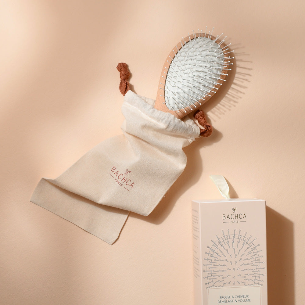 Brosse Bachca démêlage et volume en bois naturel et blanc, présentée avec son pochon en coton et son étui rose poudré.
