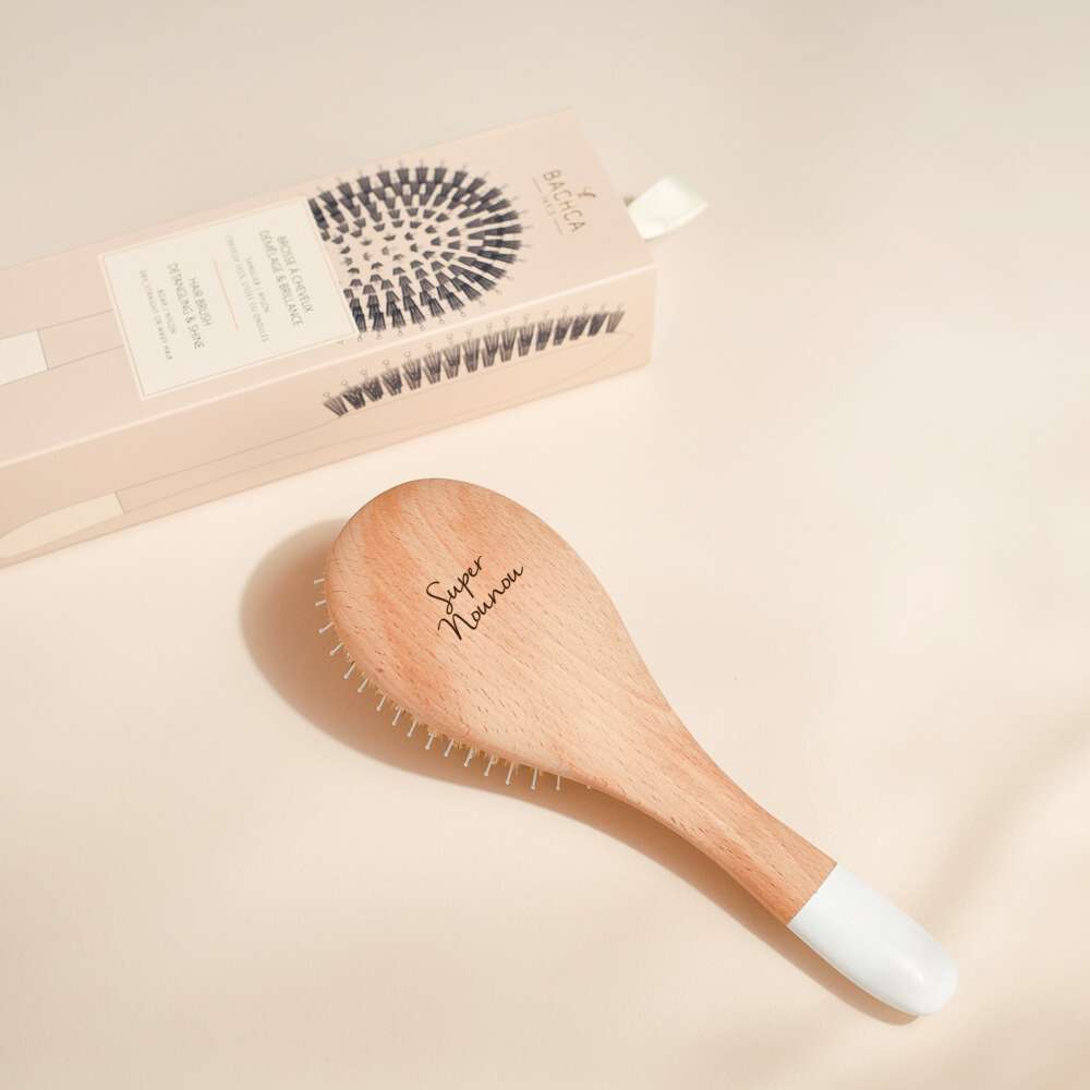 Brosse à cheveux Bachca en bois gravée "Super Nounou", posée devant sa boîte beige illustrée.