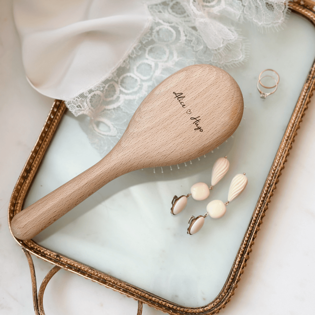 Brosse à cheveux fabriquée en France, en bois personnalisée avec gravure "Alice ♥ Hugo" de la marque Bachca, posée sur un plateau doré avec bijoux et dentelle blanche.
