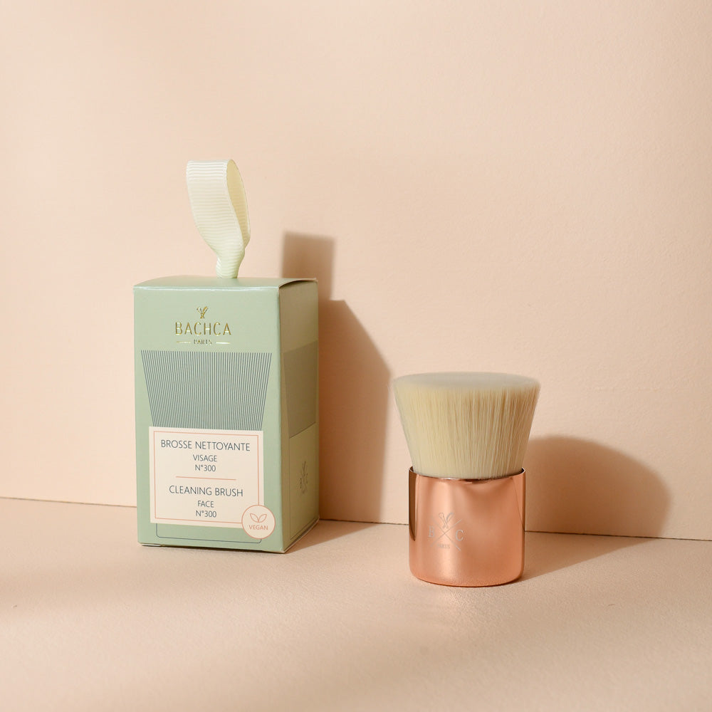 Brosse nettoyante visage Bachca avec manche cuivré et poils doux, présentée à côté de son emballage vert pastel, photographiée sur une surface beige.