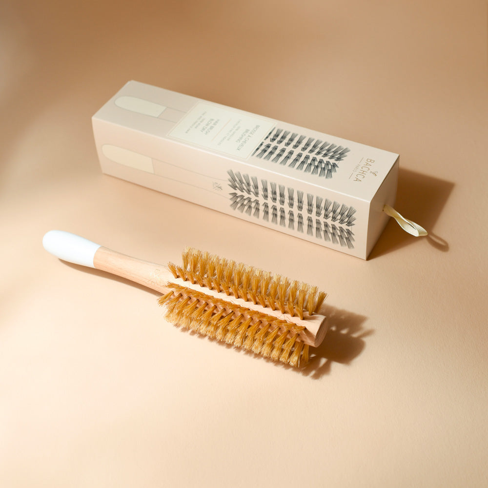 Une brosse ronde de brushing Bachca en bois clair et manche blanc est posée sur un fond beige lumineux, accompagnée de son étui cartonné assorti. Les poils naturels de la brosse se détachent avec douceur dans une mise en scène épurée et élégante.