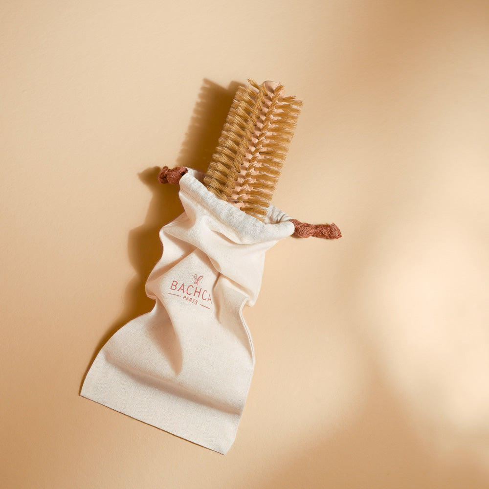 Une élégante brosse ronde de brushing en bois clair et poils naturels est présentée dans sa pochette en coton crème, siglée Bachca Paris. Posée sur un fond beige chaleureux, la mise en scène met en valeur son raffinement et son côté pratique, idéal pour protéger et transporter la brosse avec soin.