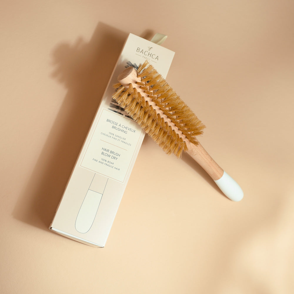 Une brosse ronde de brushing Bachca au manche en bois clair et extrémité blanche est posée sur son étui cartonné beige. Ses poils naturels de sanglier, parfaitement disposés, se détachent avec douceur dans une ambiance lumineuse et minimaliste.