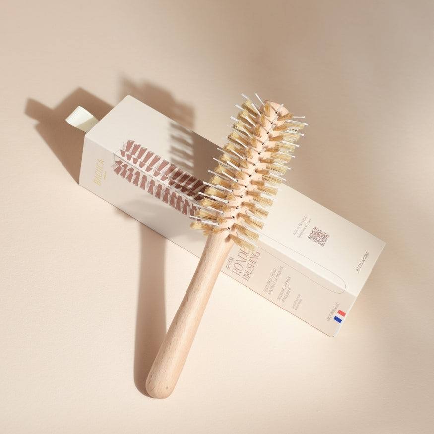 Brosse ronde brushing Bachca posée en biais sur son étui, montrant son manche en bois de hêtre brut et ses poils en nylon et soie de sanglier, éclairée par une lumière douce sur fond beige.