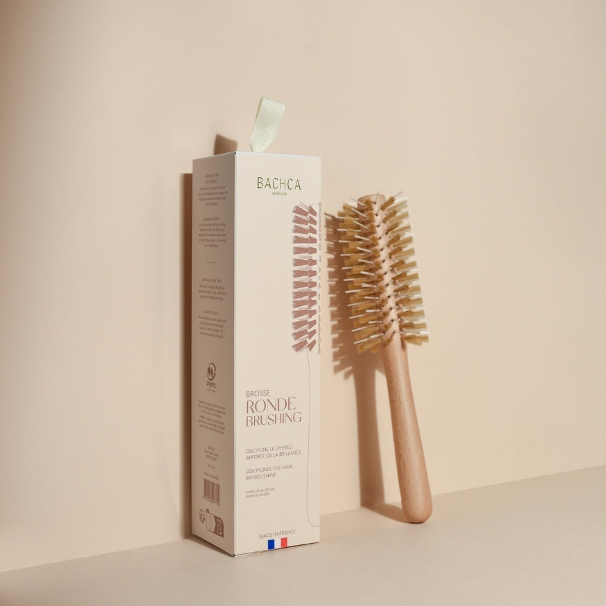 Brosse ronde brushing Bachca en bois clair, positionnée verticalement à côté de son étui beige au design épuré, soulignée par une lumière douce sur fond ton sur ton.