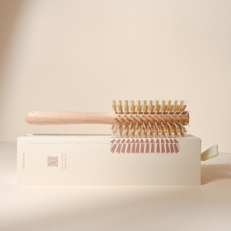 Brosse ronde de brushing Bachca en bois clair posée horizontalement sur sa boîte crème, vue de profil, avec une belle mise en valeur des poils mixtes blancs et beiges et de son packaging illustré.
