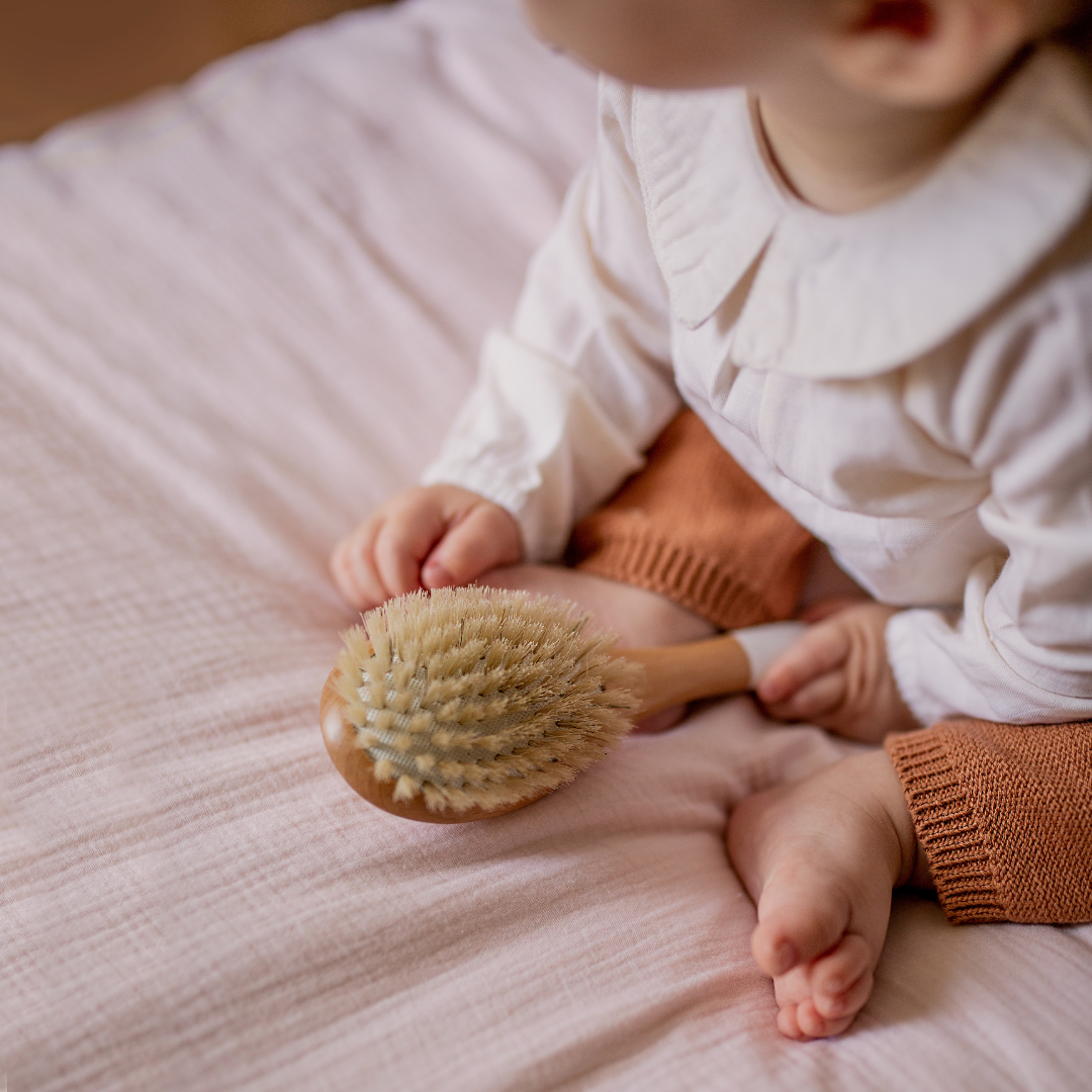 Bébé assis sur un plaid rose pâle tenant une brosse Bachca en poils de sanglier, adaptée aux cheveux fins et au cuir chevelu sensible.