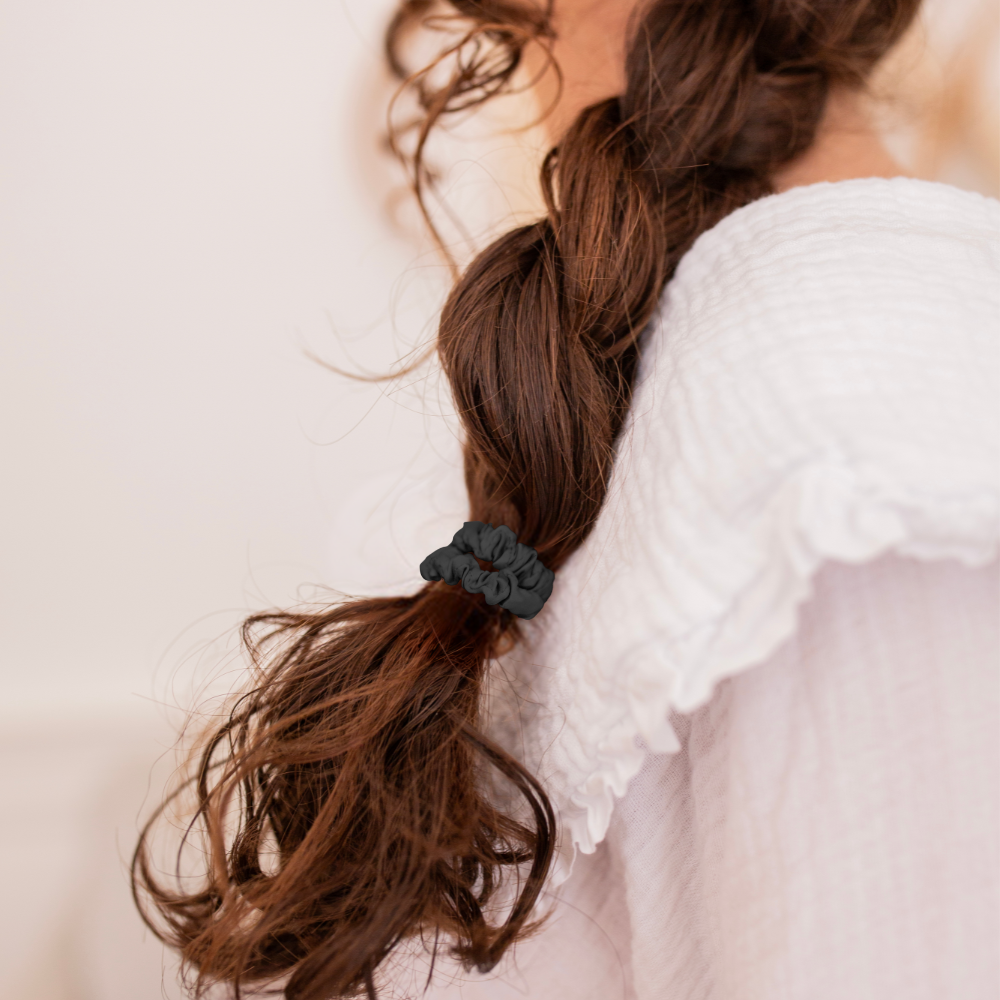 Chouchou noir en soie Bachca attachant une longue tresse souple sur une chevelure brune, porté par une jeune fille vêtue d’une blouse blanche à col volanté.